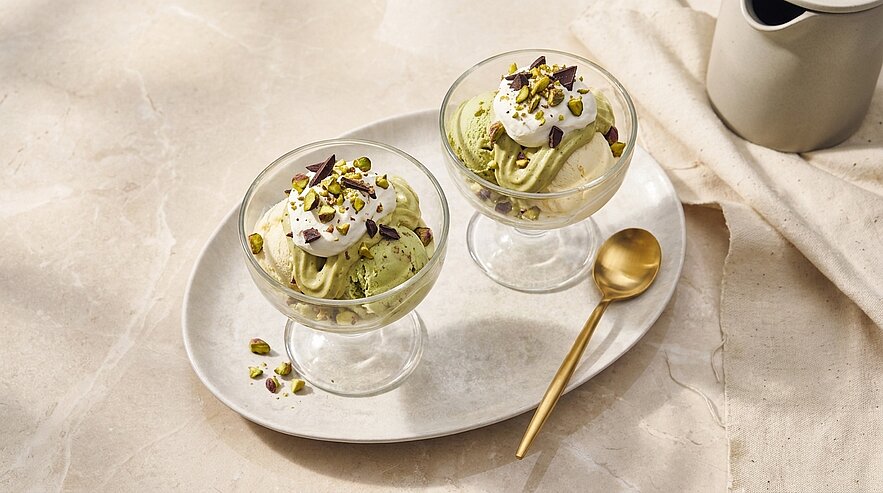 Zwei Glasdessertschalen mit Pistazieneis, jeweils mit Schlagsahne und gehackten Pistazien garniert, auf einer ovalen Platte neben einem goldenen Dessertlöffel.