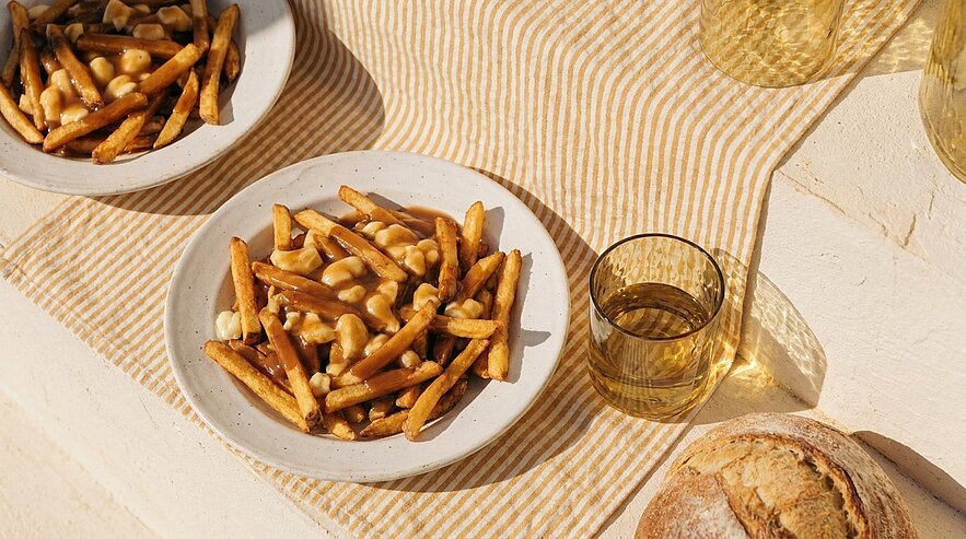 Teller mit Pommes frites, brauner Sauce und Käsebruch (Poutine) auf gestreifter Tischdecke.