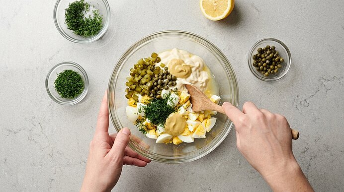 Zwei Hände verrühren gehacktes Ei, Mayonnaise, Cornichons, Kapern, Dill und Senf mit einem Holzlöffel in einer Schüssel.