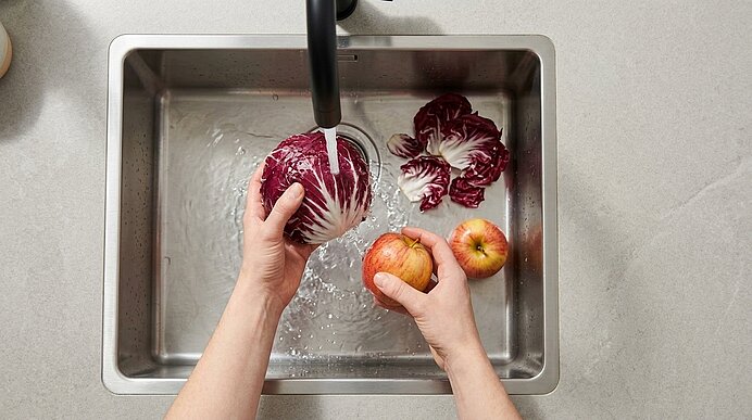 Zwei Hände waschen einen Kopf Radicchio und einen Apfel unter fließendem Wasser in einem Edelstahl-Spülbecken.