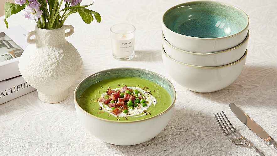 Eine Schüssel mit grüner Suppe, garniert mit Erbsen und gewürfeltem Schinken, steht auf einem Tisch. Daneben sind drei weitere Schalen gestapelt. Im Hintergrund befindet sich eine Vase mit Blumen und eine Kerze.