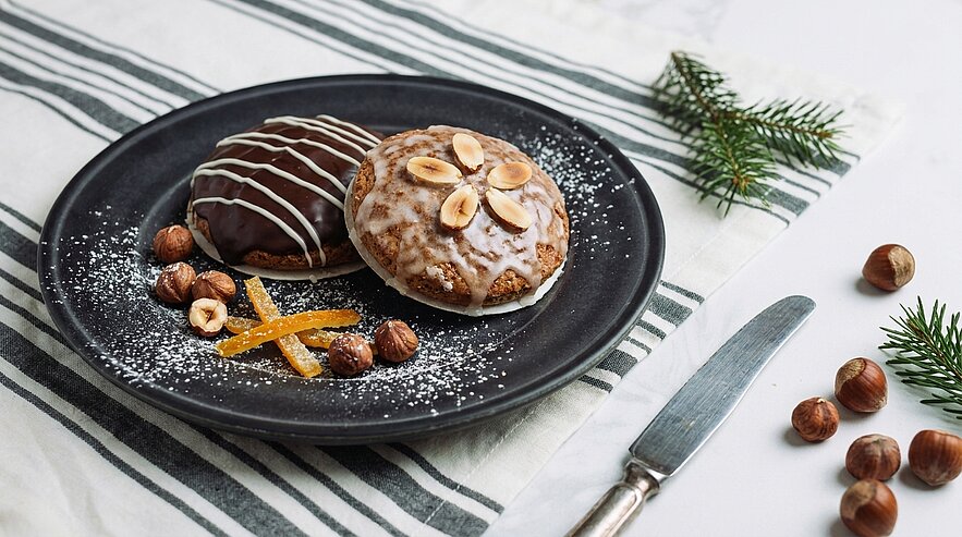Ein schwarzer Teller mit zwei Plätzchen: eines mit Schokolade und dekoriert mit Mandelscheiben, das andere mit Zuckerguss und ebenfalls Mandeln. Um die Plätzchen herum liegen gehackte Haselnüsse und kandierte Orangenschale, begleitet von einem kleinen Tannenzweig.