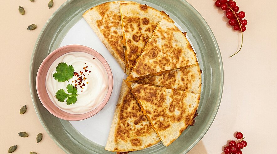 Auf einem Teller acht goldbraune Quesadilla-Dreiecke, halbkreisförmig angeordnet, daneben ein kleines Schälchen mit weißer Dip-Sauce, garniert mit Korianderblättern und Chiliflocken.