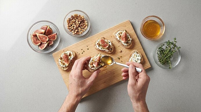 Zwei Hände beträufeln belegte Ziegenkäse-Feigen-Crostini auf einem Holzbrett mit Honig.