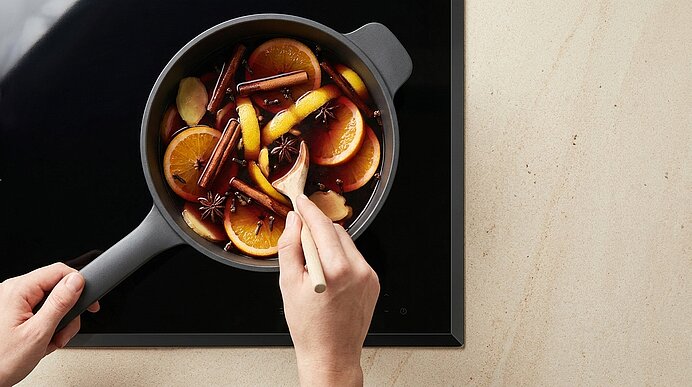 Zwei Hände rühren in einem Topf Glühwein mit Orangenscheiben, Zitronenscheiben, Zimtstangen und Sternanis auf dem Herd.