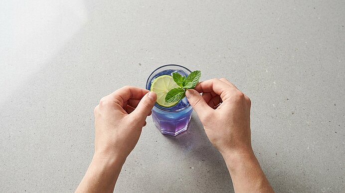 Zwei Hände legen einen Minzzweig auf ein blau-violettes Cocktailglas, das oben mit einer Limettenscheibe garniert ist.