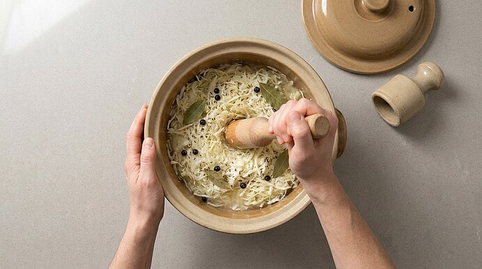 Zwei Hände stampfen gehobelten Weißkohl (Sauerkraut) mit einem Holzstampfer in einem Keramiktopf; im Kraut sind Lorbeerblätter und Pfefferkörner sichtbar.