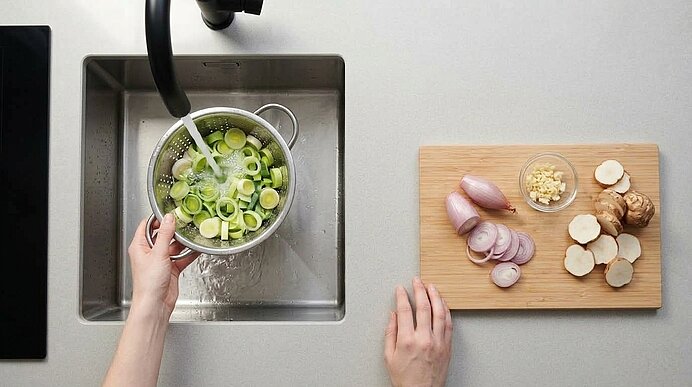 Lauchringe werden im Sieb unter fließendem Wasser gewaschen. Daneben liegen Schalotten, Knoblauch und Topinambur.