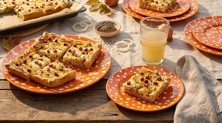 Quadratische Stücke einer herzhaften Tarte mit Speck und Zwiebeln auf orange-weiß gepunkteten Tellern auf rustikalem Holztisch, daneben ein Glas hellen Safts — herbstliche Tischszenerie.
