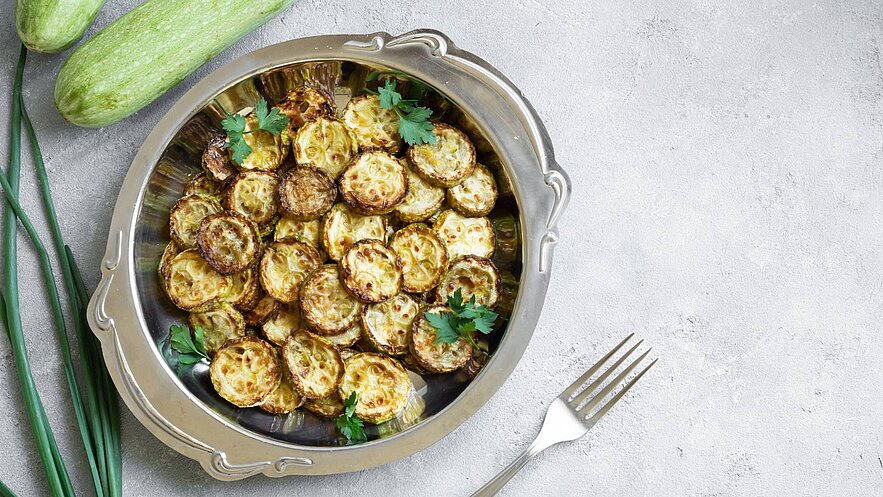 Ein silbernes Serviergericht mit goldbraun gebratenen Zucchinischeiben, garniert mit frischem Koriander. Im Hintergrund sind frische Zucchini und grüne Zwiebeln zu sehen. Der Untergrund ist hell und strukturiert.