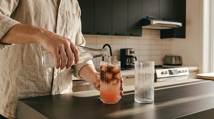 Hände einer Person gießen ein rosafarbenes, mit Eis gefülltes Getränk aus einer Glasflasche in ein hohes Glas auf einer Küchenarbeitsplatte.