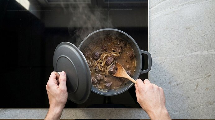 Zwei Hände heben den Deckel eines Kochtopfs und rühren mit einem Holzlöffel ein köchelndes Fleischgulasch.
