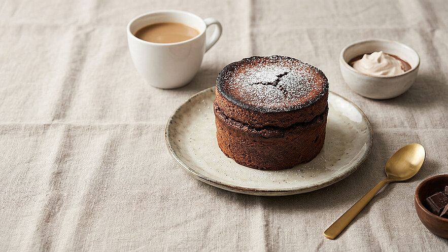 Ein großer, schokoladiger Kuchen auf einem Teller, bestäubt mit Puderzucker, wird von einer Tasse Kaffee und einer Schüssel mit Sahne umgeben. Ein goldener Löffel liegt neben dem Teller, während eine Schüssel mit Schokolade im Hintergrund sichtbar ist.