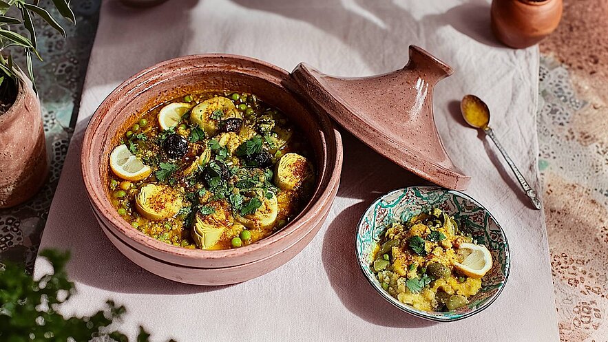 Ein rustikaler, terrakottafarbener Tajine-Topf gefüllt mit einem bunten Gericht aus Erbsen, Zitronenscheiben und frischem Koriander. Daneben befindet sich eine kleine Schüssel mit einer ähnlichen Mischung. Die Speisen wirken einladend und aromatisch angerichtet.
