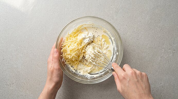 Zwei Hände verrühren geriebenen Bergkäse, Frischkäse, Sahne und Pfeffer mit einem Schneebesen in einer Glasschüssel.