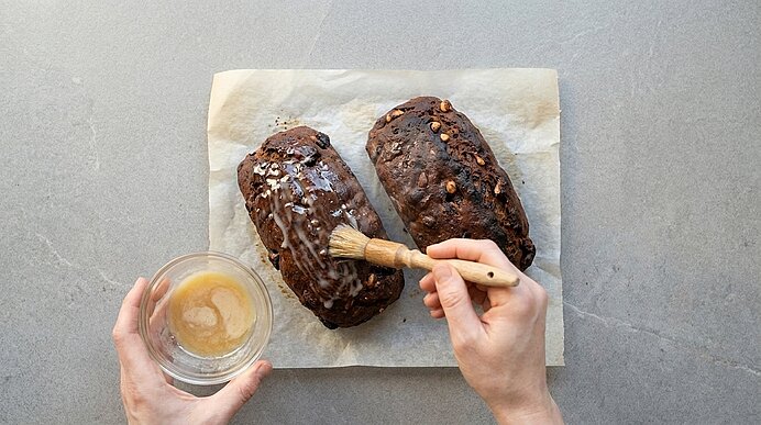 Zwei schokoladige Laibe mit Nüssen auf Backpapier; Hände bestreichen einen Laib mit Glasur aus einer kleinen Schüssel.