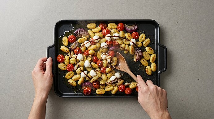 Backblech mit gerösteten Gnocchi, Kirschtomaten, roten Zwiebeln und Mozzarellakugeln, mit Kräutern und Öl, von einer Hand mit Holzlöffel durchmischt.