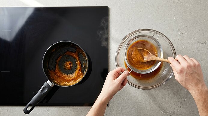 Hände schöpfen mit einem Holzlöffel dicke braune Sauce aus einer Glasschüssel und geben sie in eine kleine Pfanne auf dem Herd.