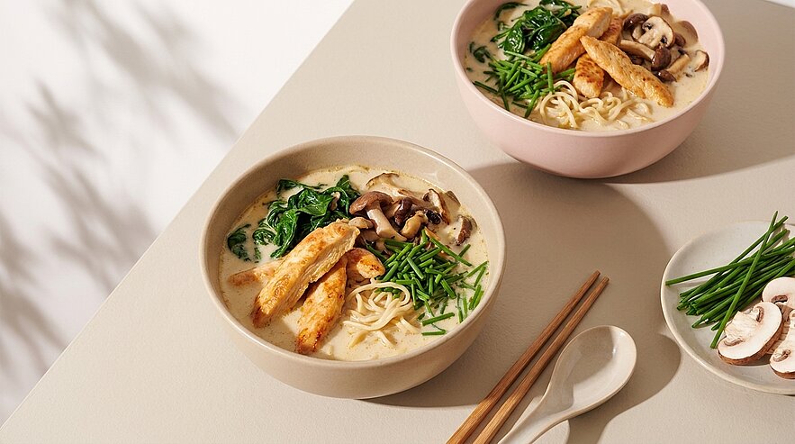 Zwei Schalen mit nudelbasierter Suppe sind auf einem Tisch platziert. Die erste Schale enthält Stücke von gebratenem Hähnchen, Spinat und Pilzen. Die zweite Schale zeigt ähnliche Zutaten. Neben den Schalen liegen Essstäbchen und ein kleiner Teller mit frischen Kräutern und weiteren Pilzen.