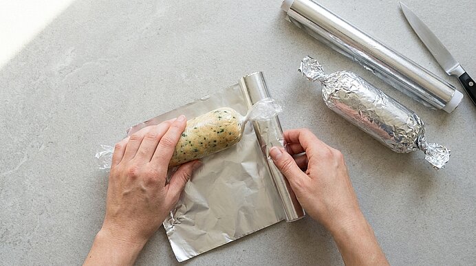 Hände wickeln eine Kräuterbutter- oder Teigrolle in Frischhaltefolie auf Aluminiumfolie; daneben liegt eine in Alufolie verpackte Rolle und ein Messer.