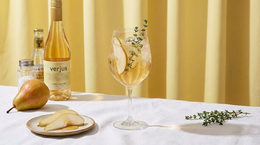 Weinglas mit hellem Verjus-Cocktail, Eis, Birnenscheiben und Thymian; daneben eine Flasche "verjus", eine ganze Birne, ein Teller mit Birnenscheiben und ein Thymianzweig auf weißer Tischdecke.
