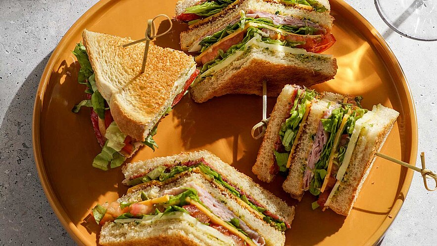 Ein Teller mit mehreren Sandwiches, die in Stücke geschnitten sind. Die Sandwiches sind mit verschiedenen Zutaten gefüllt, darunter Salat, Tomaten und Aufschnitt. Die Brotscheiben sind knusprig gebräunt. Ein Zahnstocher hält einige der Stücke zusammen.