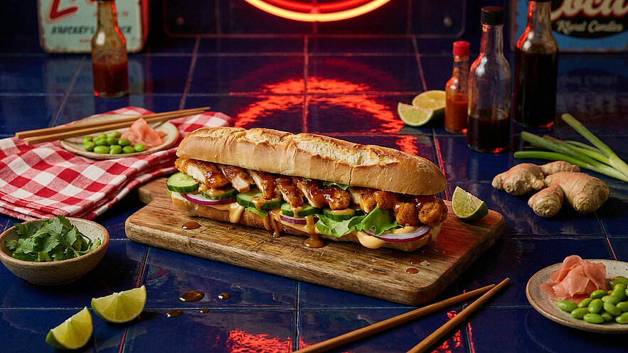 Ein großer Sandwich auf einem Holzbrett, gefüllt mit Hähnchen, Gurken, Salat und Sauce. Daneben kleine Schalen mit Ingwer und Edamame, sowie Limettenscheiben und eine kariert rot-weiße Serviette. Hintergrund mit einer Neonleuchte und verschiedenen Flaschen.
