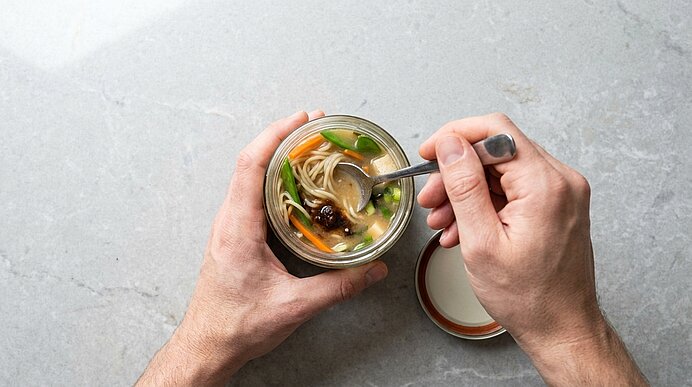 Hände halten ein Glas mit Nudelsuppe (Brühe und Gemüse) und rühren mit einem Löffel; der Glasdeckel liegt daneben.