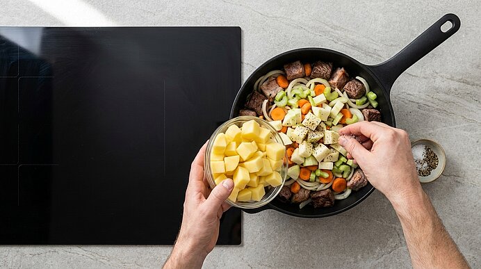 Hände geben gewürfelte Kartoffeln in eine Pfanne mit Fleisch und Gemüse (Karotten, Sellerie) auf dem Herd.
