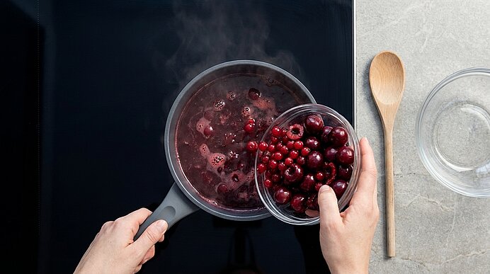 Hände füllen frische Cranberries aus einer Glasschüssel in einen dampfenden Topf mit roter Fruchtmischung auf dem Herd.