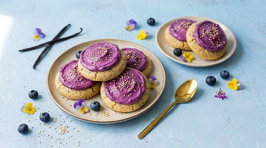 Zwei Teller mit runden Keksen, jeweils mit violettem Frosting und Sesam bestreut; dekoriert mit Blaubeeren und essbaren Blüten, daneben ein goldener Löffel.
