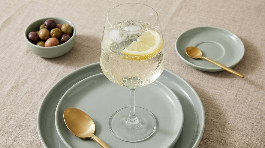 Ein stilvoll gedeckter Tisch mit einem Glas Sprudelwasser, das mit einer Zitronenscheibe dekoriert ist. Daneben liegen große und kleine graue Teller sowie eine kleine Schüssel mit einer Vielzahl von Oliven. Eine goldene Löffel liegt bereit.