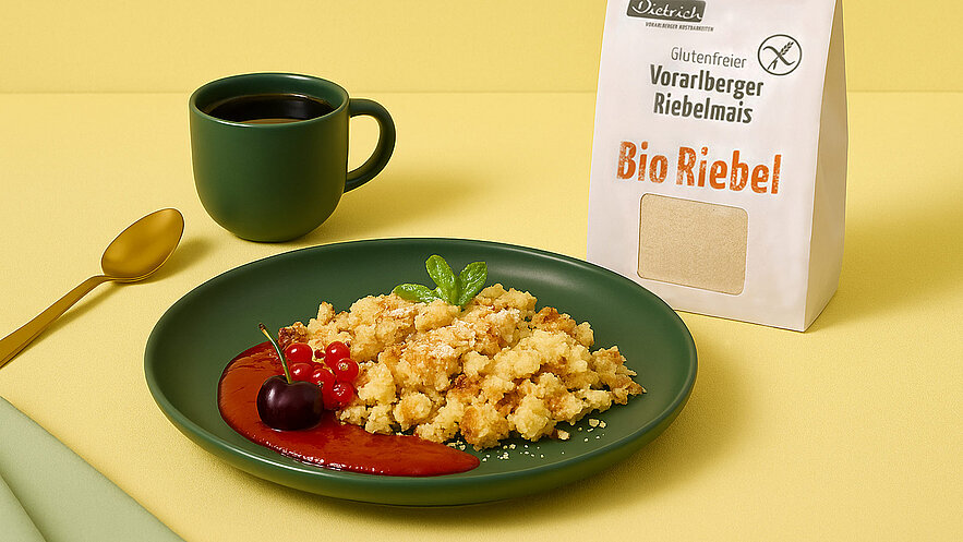 Auf dem Bild sind drei knusprige Riebels aus Vorarlberger Riebelmais auf einem grünen Teller zu sehen. Daneben stehen eine goldene Löffel, eine Tasse Kaffee und ein verpackter Riebelmais auf einem gelben Hintergrund.