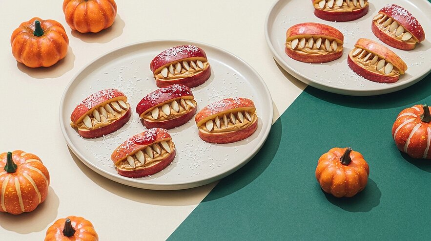 Rote Kekse mit Vanillecreme und Mandeln als spitze Zähne, arrangiert wie kleine Münder auf Tellern, umgeben von Mini-Kürbissen als Halloween-Dekor.
