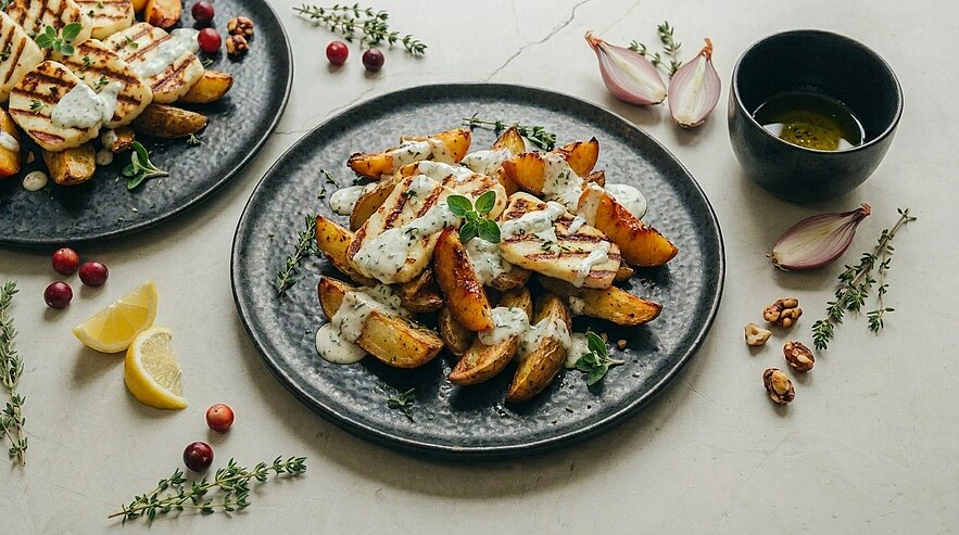 Dunkler Teller mit goldbraunen Kartoffelspalten, beträufelt mit cremiger Kräutersauce und garniert mit frischen Kräutern.