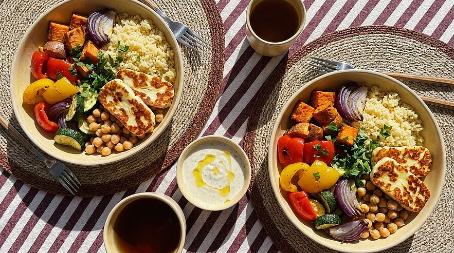 Zwei bunte Bowls mit Couscous, geröstetem Gemüse (Paprika, Zwiebel, Süßkartoffel), Kichererbsen, gegrilltem Halloumi und frischen Kräutern, dazu eine kleine Schale Joghurt-Dip und zwei Getränke auf gestreifter Tischdecke.