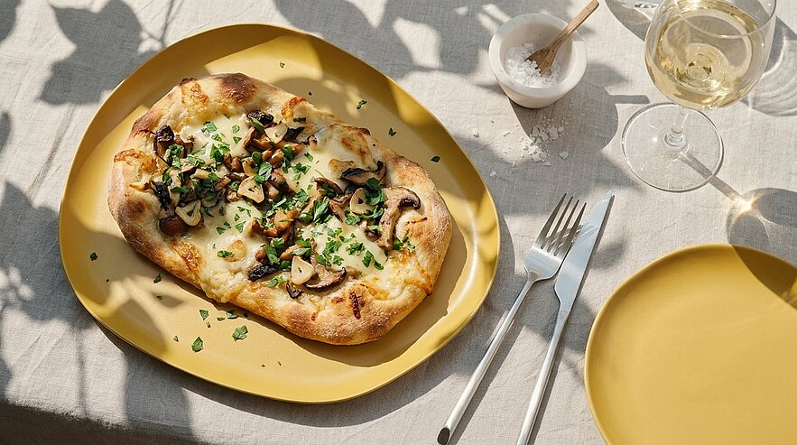 Ein goldener Teller mit einer belegten Pizza, die mit geschmolzenem Käse, Pilzen und frischen Kräutern belegt ist. Daneben ein Glas Weißwein und eine kleine Schale mit Salz. Der Hintergrund ist ein gedeckter Tisch mit einer hellen Tischdecke.