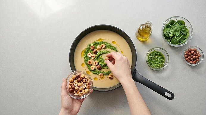 Zwei Hände garnieren helle Topinambursuppe in einer mattschwarzen Stielpfanne mit grünem Pesto und Haselnüssen.