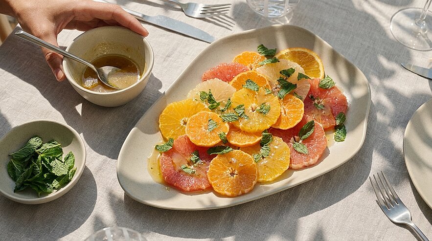Teller mit aufgeschnittenen Orangen- und Grapefruitscheiben, garniert mit frischer Minze; daneben ein Schälchen mit Dressing, in das eine Hand einen Löffel hält.