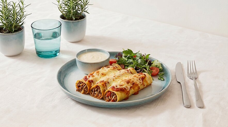 Drei überbackene, gefüllte Pastarollen (Cannelloni) auf einem Teller mit kleinem Dip und gemischtem Salat; Besteck und Glas Wasser daneben.