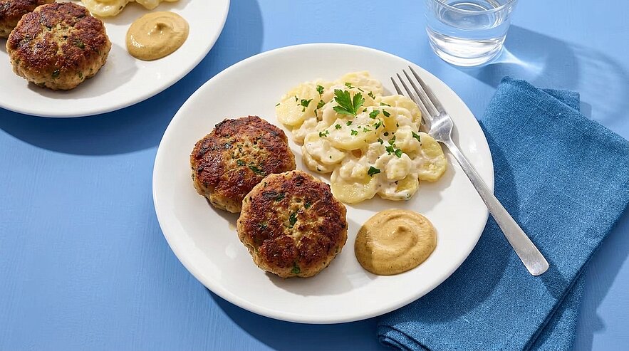 Weißer Teller mit zwei gebratenen Frikadellen, Kartoffelsalat mit Petersilie und einem Klecks Senf; Gabel und blaues Stofftuch daneben.