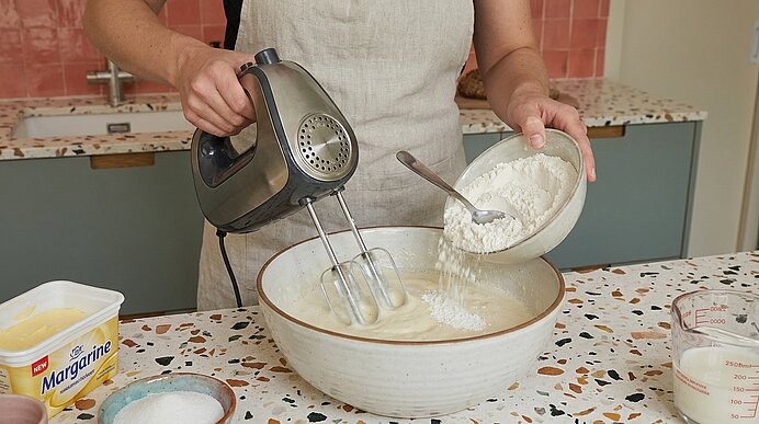 Person in Küchenschürze rührt mit elektrischem Handmixer in einer großen Schüssel, während sie Mehl aus einer kleinen Schüssel in den Teig schüttet.