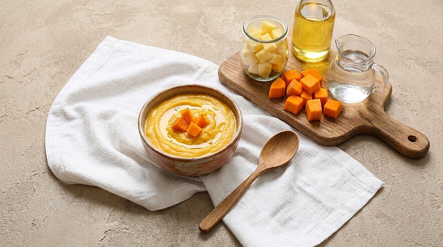 Schale mit cremiger Kürbissuppe, garniert mit Kürbiswürfeln; daneben Holzlöffel und Brett mit zusätzlichen Kürbiswürfeln sowie Öl- und Wassergläser auf weißer Serviette.