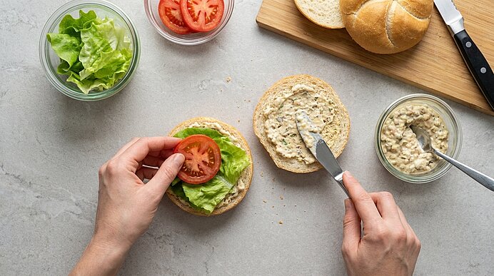 Zwei Hände bestreichen Brötchenhälften mit Remoulade und belegen eine Hälfte mit Salat und einer Tomatenscheibe.