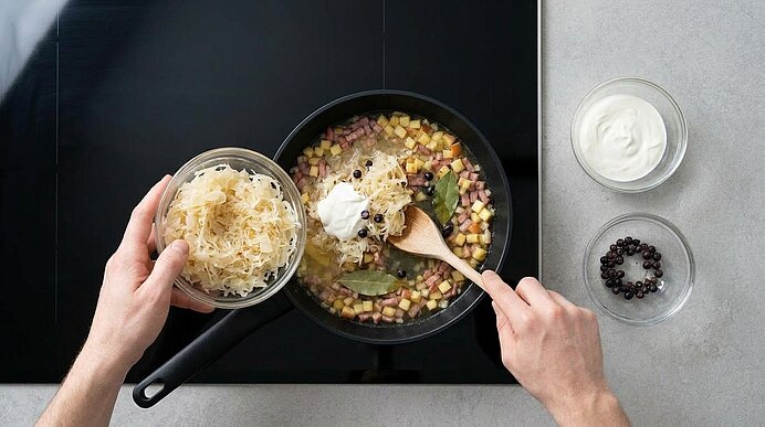 Zwei Hände geben Sauerkraut in eine Pfanne mit Zwiebel-, Apfel- und Schinkenwürfeln, Brühe, Schmand und Gewürzen.