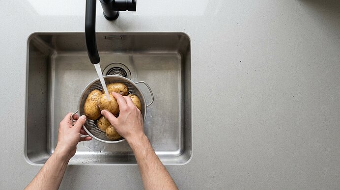 Zwei Hände waschen Kartoffeln in einem Sieb unter fließendem Wasser in einer Küchenspüle.