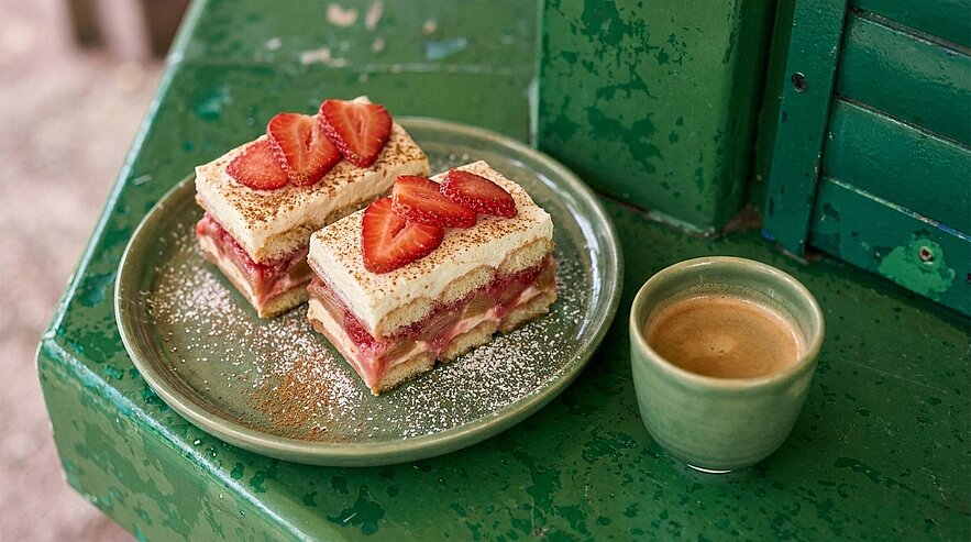 Zwei Stücke geschichteter Erdbeer-Sahne-Kuchen mit Puderzucker und frischen Erdbeerscheiben auf einem grünen Teller, daneben eine kleine Tasse Espresso.