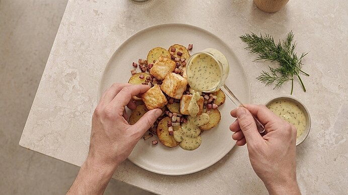 Zwei Hände garnieren einen Teller mit gebratenen Kartoffelscheiben, knusprigen Würfeln, kleinen Speckstückchen und einer cremigen Dill-Senfsauce.