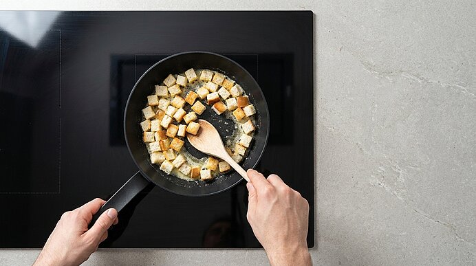 Hände rühren in einer Pfanne auf einem Kochfeld geröstete Brotwürfel (Croutons) mit einem Holzlöffel.