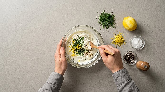 Zwei Hände rühren gehackten Dill, Zitronenabrieb und Pfeffer in einer Glasschüssel in eine helle Rahmcreme.
